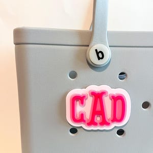 Puede incluir: Un bolso tote gris claro con un asa azul claro y un charm rosa y blanco que dice "CAD". El charm est&aacute; sujeto a la bolsa con un bot&oacute;n blanco que tiene una "b" negra.