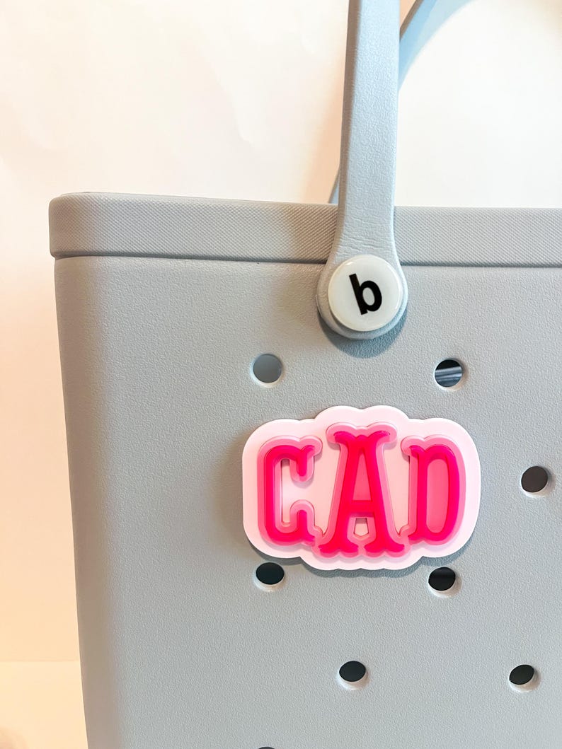 Puede incluir: Un bolso tote gris claro con un asa azul claro y un charm rosa y blanco que dice "CAD". El charm est&aacute; sujeto a la bolsa con un bot&oacute;n blanco que tiene una "b" negra.