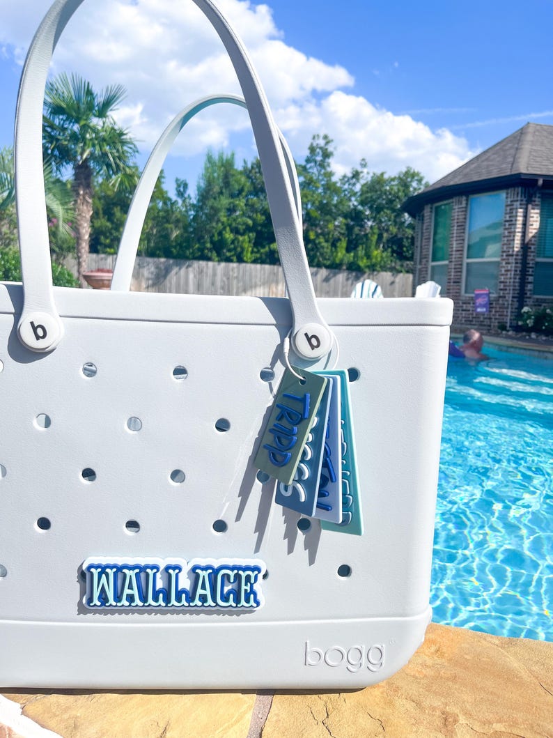 Puede incluir: Un bolso tote gris claro con asas blancas y el nombre "WALLACE" en letras azules. El bolso tiene una serie de peque&ntilde;os agujeros y una etiqueta que dice "TRIPP". El bolso est&aacute; cerca de una piscina en un d&iacute;a soleado.