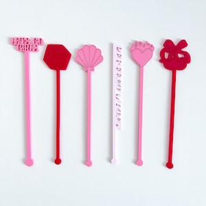 May include: A collection of six drink stirrers in shades of pink and red, arranged against a white background. The stirrers feature various shapes, including a hexagon, seashell, heart, and bow. One stirrer has the text "LET'S GO GIRLS".