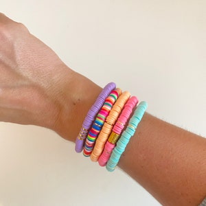 May include: A set of five colorful beaded bracelets. The bracelets are stacked on a person's wrist. The bracelets are made of small, round beads in shades of purple, pink, orange, and blue. Some of the beads have a gold accent.