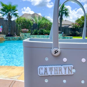 Puede incluir: Un bolso tote gris claro con asas blancas y el nombre "CATHRYN" en letras plateadas con purpurina. El bolso tiene varios recortes circulares blancos, cerca de una piscina.