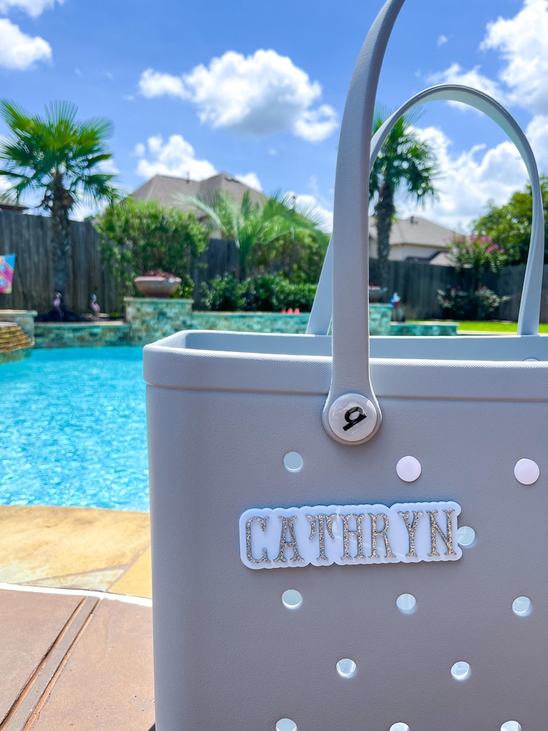 Puede incluir: Un bolso tote gris claro con asas blancas y el nombre "CATHRYN" en letras plateadas con purpurina. El bolso tiene varios recortes circulares blancos, cerca de una piscina.