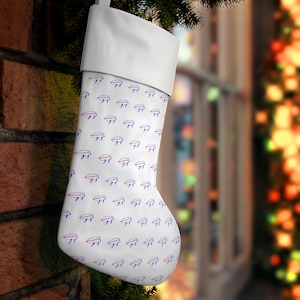 Può includere: Calza di Natale bianca con un motivo ripetuto di loghi di testa di bufalo blu e rossi. La calza ha un polsino bianco ed è appesa a un muro di mattoni. Una decorazione festiva.