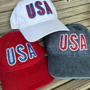 May include: Three baseball caps in white, red, and denim blue. Each cap features the word "USA" in a bold, outlined font with red and blue accents. The caps are arranged on a wooden surface.
