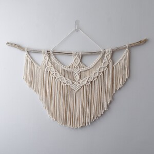 May include: Cream-coloured macrame wall hanging. The textile art piece features a detailed geometric design with a fringe at the bottom. It is suspended from a natural wooden branch, creating a bohemian aesthetic. The wall decor is hung against a plain white wall.