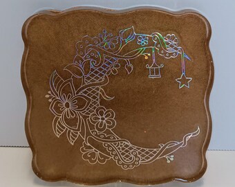 May include: A brown, square resin tray with a scalloped edge. The tray features a white, floral crescent moon design with a star and lantern. The design has a holographic, iridescent sheen.