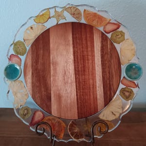 May include: A round wooden serving board with a clear resin edge inlaid with dried fruit slices. The board has a light brown and reddish-brown striped pattern. Two turquoise ceramic knobs are on the sides.