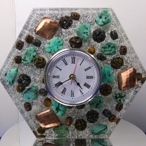 May include: A decorative hexagonal clock with a silver glitter background. The clock face is white with black Roman numerals and a red second hand. Turquoise stones, copper-colored squares, and bronze beads are embedded in the clock's face.
