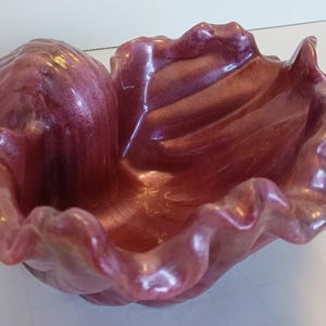 May include: A decorative bowl in shades of burgundy and rose with an iridescent finish. The bowl is shaped like a seashell with a ruffled edge. The surface has a glossy finish, reflecting light. The bowl is designed for decorative purposes.