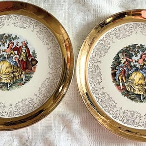 May include: Two decorative plates with gold rims and a central image of a couple in period clothing dancing. The plates have a cream-colored background with a gold floral pattern around the central image.