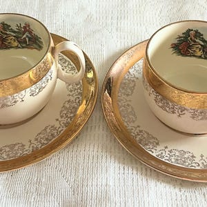 May include: Two vintage teacups and saucers. The cups and saucers are cream-colored with gold trim and a gold band around the cup's rim. Each cup has a small image of a couple in a garden. The saucers have a gold rim and a decorative pattern.