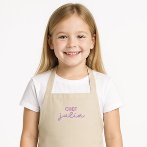 May include: A young girl wearing a tan apron with a large front pocket. The apron has the word "CHEF" above the name "julia" in a purple script. The girl is wearing a white t-shirt.