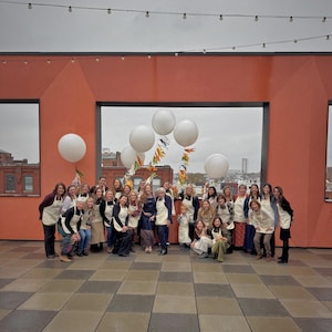 Puede incluir: Un grupo de personas, en su mayor&iacute;a mujeres, posan para una foto en una azotea. Llevan delantales y est&aacute;n rodeadas de globos blancos y guirnaldas decorativas. El fondo incluye una pared naranja y un paisaje urbano.