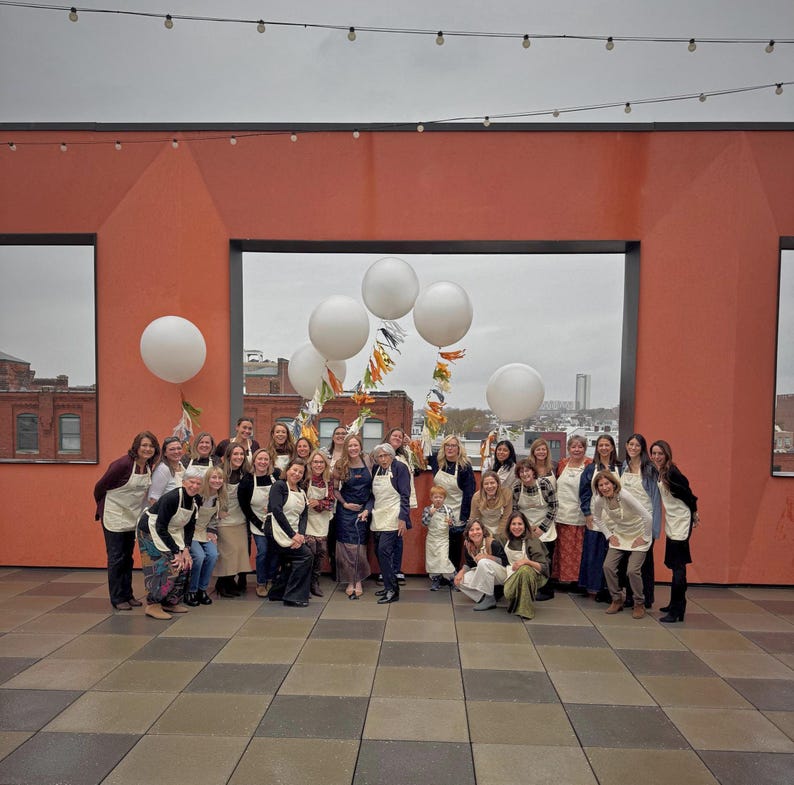 Puede incluir: Un grupo de personas, en su mayor&iacute;a mujeres, posan para una foto en una azotea. Llevan delantales y est&aacute;n rodeadas de globos blancos y guirnaldas decorativas. El fondo incluye una pared naranja y un paisaje urbano.