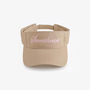 Pode incluir: Viseira bege com a palavra "Sweetheart" bordada em rosa. A viseira tem uma aba curva e uma alça ajustável. Feita de um material macio e durável, para proteção solar.
