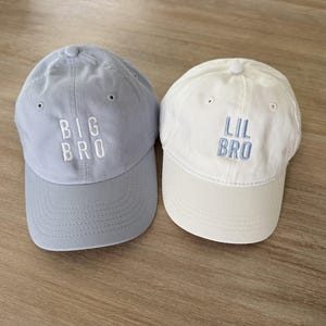 Puede incluir: Se muestran dos gorras de b&eacute;isbol, una azul claro y otra blanca. La gorra azul claro tiene bordado "BIG BRO" en blanco, mientras que la gorra blanca tiene bordado "LIL BRO" en azul claro. Ambas gorras tienen una visera curva.