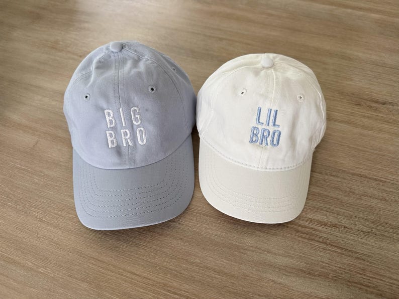 Puede incluir: Se muestran dos gorras de b&eacute;isbol, una azul claro y otra blanca. La gorra azul claro tiene bordado "BIG BRO" en blanco, mientras que la gorra blanca tiene bordado "LIL BRO" en azul claro. Ambas gorras tienen una visera curva.