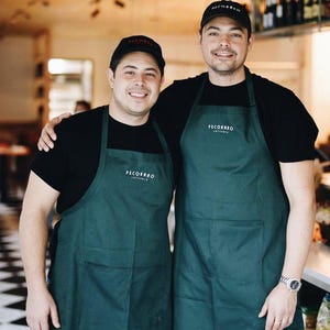 Puede incluir: Dos personas con camisetas negras, delantales verde oscuro y gorras de b&eacute;isbol negras con el texto "PECORARO" en los delantales y gorras. Est&aacute;n sonriendo en un restaurante.