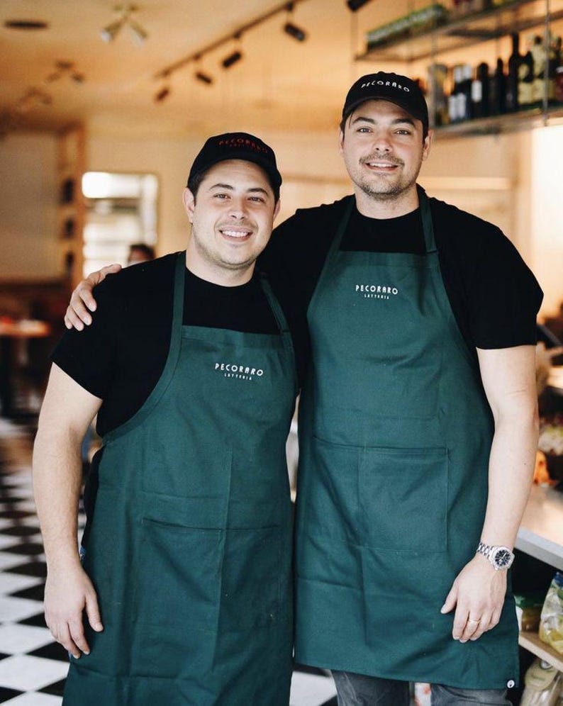 Puede incluir: Dos personas con camisetas negras, delantales verde oscuro y gorras de b&eacute;isbol negras con el texto "PECORARO" en los delantales y gorras. Est&aacute;n sonriendo en un restaurante.