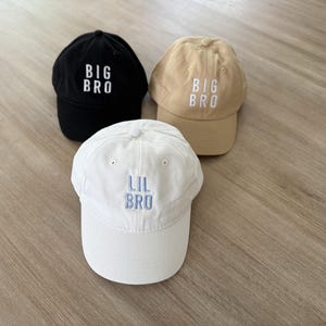 Puede incluir: Tres gorras de b&eacute;isbol en negro, beige y blanco. Las gorras negras y beige tienen "BIG BRO" bordado en blanco, mientras que la gorra blanca tiene "LIL BRO" bordado en azul. Las gorras est&aacute;n hechas de tela suave.