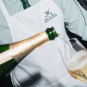 Puede incluir: Un delantal blanco con el texto "THE GLOBAL AMBASSADOR" y un logotipo de golf se usa sobre una camisa y corbata a rayas verdes. Se vierte champ&aacute;n de una botella a una copa.