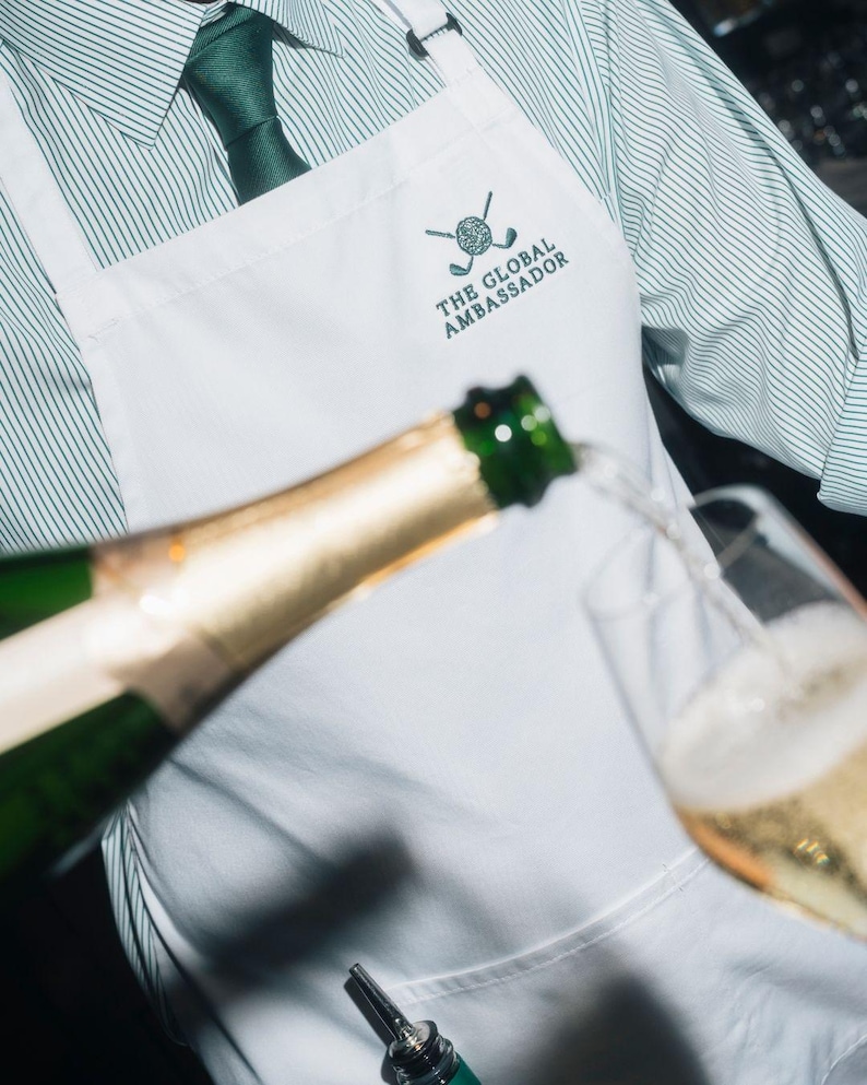 Puede incluir: Un delantal blanco con el texto "THE GLOBAL AMBASSADOR" y un logotipo de golf se usa sobre una camisa y corbata a rayas verdes. Se vierte champ&aacute;n de una botella a una copa.
