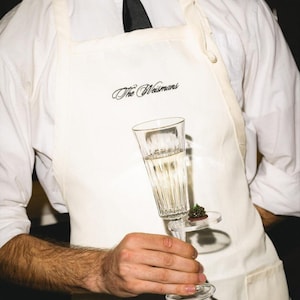 Puede incluir: Una copa de champ&aacute;n transparente con un peque&ntilde;o aperitivo en un plato transparente es sostenida por una persona que lleva un delantal blanco con el texto "The Weismans" en escritura negra. La persona tambi&eacute;n lleva una camisa blanca y una corbata negra.