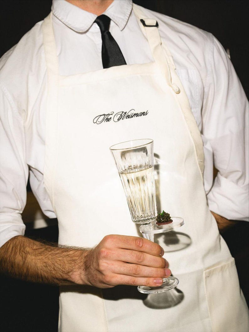 Puede incluir: Una copa de champ&aacute;n transparente con un peque&ntilde;o aperitivo en un plato transparente es sostenida por una persona que lleva un delantal blanco con el texto "The Weismans" en escritura negra. La persona tambi&eacute;n lleva una camisa blanca y una corbata negra.
