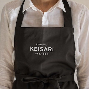 Puede incluir: Delantal negro con el texto "LEIPOMO KEISARI EST. 1990" en blanco. El delantal tiene un lazo en la cintura y correas negras. El delantal se lleva sobre una camisa blanca abotonada.