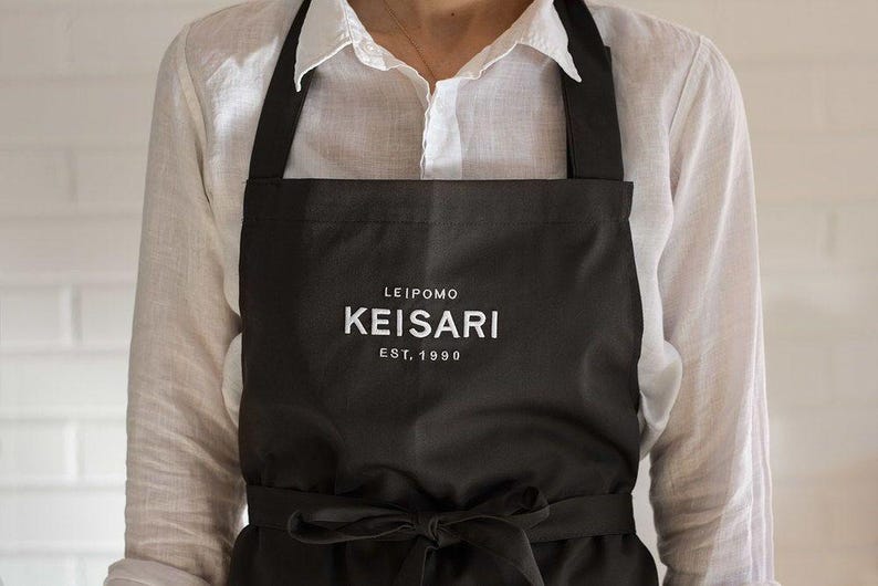 Puede incluir: Delantal negro con el texto "LEIPOMO KEISARI EST. 1990" en blanco. El delantal tiene un lazo en la cintura y correas negras. El delantal se lleva sobre una camisa blanca abotonada.