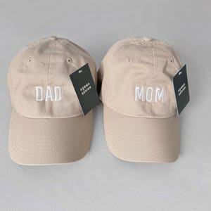 Puede incluir: Dos gorras de b&eacute;isbol beige, una bordada con "DAD" y la otra con "MOM" en blanco. Cada gorra tiene una visera curva y una etiqueta que dice "FORMA DESIGN". Las gorras son un conjunto a juego.