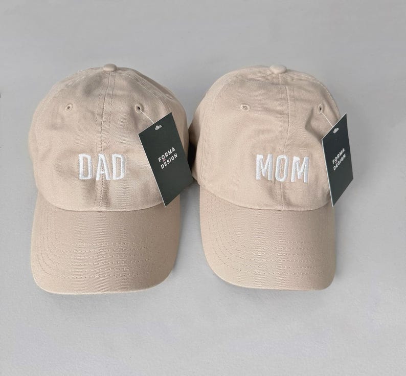 Puede incluir: Dos gorras de b&eacute;isbol beige, una bordada con "DAD" y la otra con "MOM" en blanco. Cada gorra tiene una visera curva y una etiqueta que dice "FORMA DESIGN". Las gorras son un conjunto a juego.