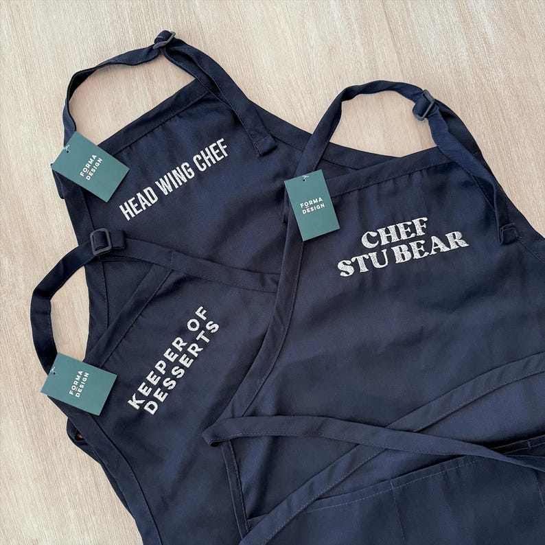 Puede incluir: Tres delantales azul marino con texto blanco. Los textos en los delantales dicen "HEAD WING CHEF", "CHEF STU BEAR" y "KEEPER OF DESSERTS". Cada delantal tiene una etiqueta que dice "FORMA DESIGN".
