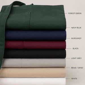 Puede incluir: Una pila de delantales plegados en varios colores, incluyendo verde bosque, azul marino, burdeos, negro, gris claro, beige/arena y blanco. Los delantales tienen un bolsillo y correas largas.