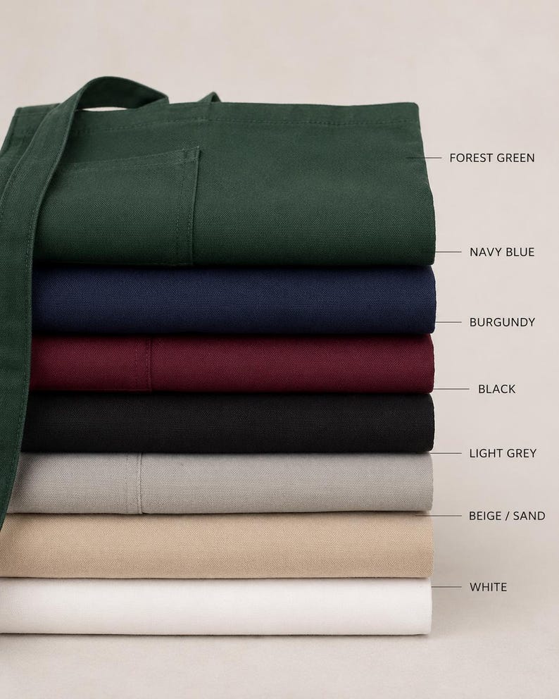 Puede incluir: Una pila de delantales plegados en varios colores, incluyendo verde bosque, azul marino, burdeos, negro, gris claro, beige/arena y blanco. Los delantales tienen un bolsillo y correas largas.