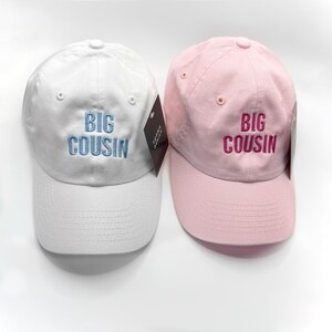 Puede incluir: Se muestran dos gorras de b&eacute;isbol, una blanca y otra rosa. La gorra blanca tiene bordado "BIG COUSIN" en azul claro, mientras que la gorra rosa tiene el mismo texto en rosa m&aacute;s oscuro. Ambas gorras tienen una visera curva y un bot&oacute;n en la parte superior.