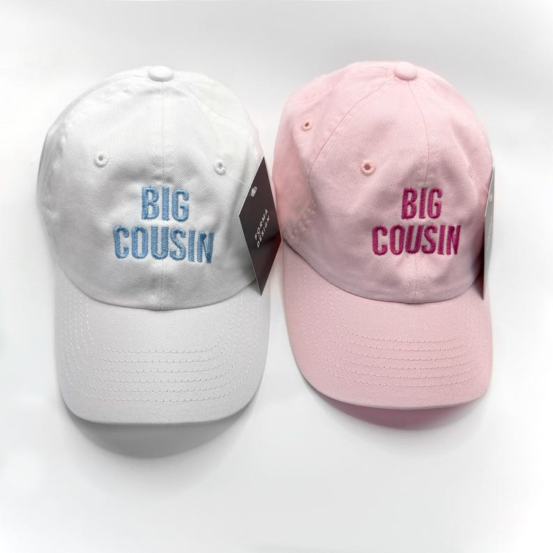 Puede incluir: Se muestran dos gorras de b&eacute;isbol, una blanca y otra rosa. La gorra blanca tiene bordado "BIG COUSIN" en azul claro, mientras que la gorra rosa tiene el mismo texto en rosa m&aacute;s oscuro. Ambas gorras tienen una visera curva y un bot&oacute;n en la parte superior.