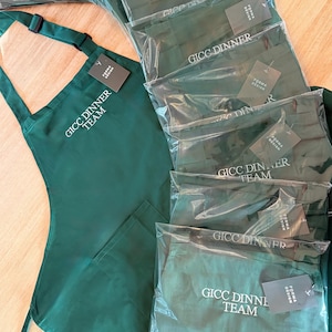 Puede incluir: Una colecci&oacute;n de delantales verde oscuro y art&iacute;culos a juego, algunos empaquetados individualmente en pl&aacute;stico transparente. Los delantales tienen el texto "GICC DINNER TEAM" impreso en blanco. Cada art&iacute;culo tiene una etiqueta "FORMA DESIGN".