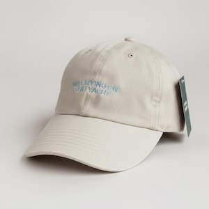 May include: A beige baseball cap with a curved brim. The cap features the light blue text "NO CRYING ON THE YACHT" embroidered on the front. A green tag hangs from the side. The cap is made of a soft material.