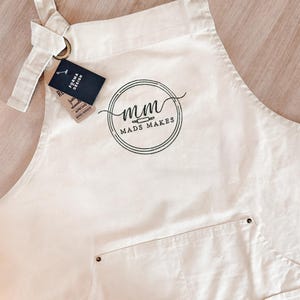 Puede incluir: Delantal color crema con un bolsillo y el texto "mads makes" en un logotipo circular. El delantal tiene una correa para el cuello y lazos en la cintura. Una etiqueta dice "FORMA DESIGN APRONS".