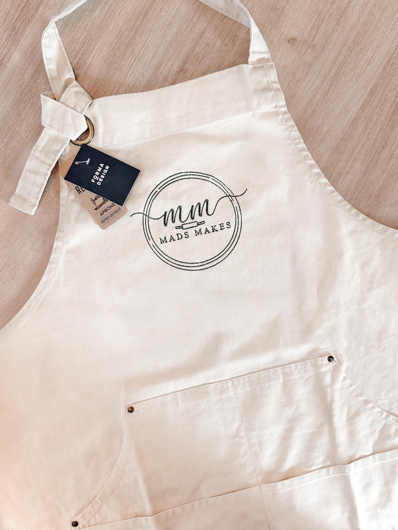 Puede incluir: Delantal color crema con un bolsillo y el texto "mads makes" en un logotipo circular. El delantal tiene una correa para el cuello y lazos en la cintura. Una etiqueta dice "FORMA DESIGN APRONS".
