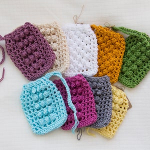 May include: Assortment of ten crocheted soap saver bags in various colors, including purple, beige, white, yellow, green, blue, pink, gray, and light yellow. Each bag has a loop for hanging.
