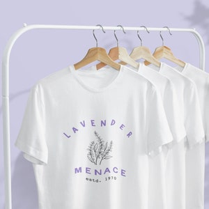 May include: Three white t-shirts hanging on wooden hangers on a white rack. The front t-shirt has a purple graphic with the text "Lavender Menace estd. 1970".