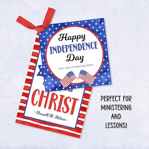May include: Two rectangular paper tags with red ribbon. The top tag features a blue background with white stars, two American flags, and the text Happy Independence Day love, your ministering sisters. The bottom tag has red stripes and text CHRIST.