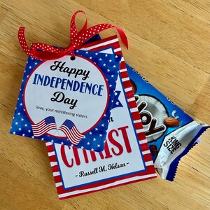 May include: A square card with a red, white, and blue star pattern, featuring the text Happy Independence Day, love, your ministering sisters, and two American flags. Below, a card reads CHRIST and Russell M. Nelson. A red polka dot ribbon is attached.