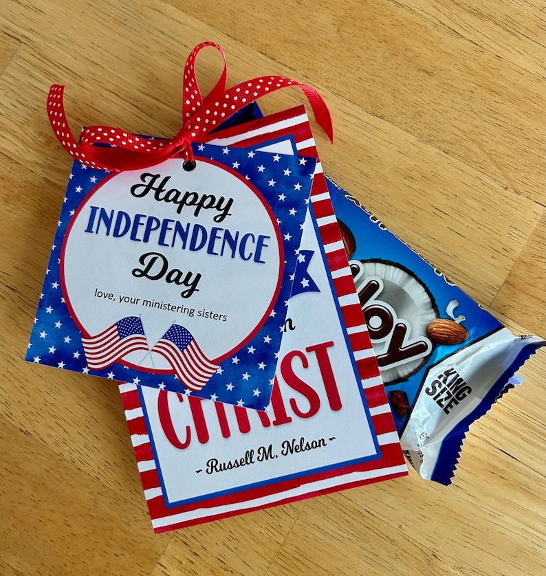 May include: A square card with a red, white, and blue star pattern, featuring the text Happy Independence Day, love, your ministering sisters, and two American flags. Below, a card reads CHRIST and Russell M. Nelson. A red polka dot ribbon is attached.