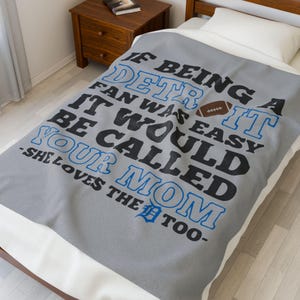 May include: A grey blanket featuring the text "IF BEING A DETROIT FAN WAS EASY IT WOULD BE CALLED YOUR MOM -SHE LOVES THE TOO-" in black and blue, with a football graphic. The blanket is on a bed.