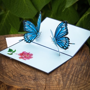 May include: A blue and white pop-up card with two butterflies and a pink flower. The card is open and the butterflies are in a 3D pop-up design.