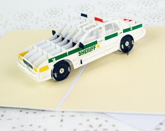 Puede incluir: Una tarjeta de felicitación pop-up de un coche de sheriff blanco y verde. La tarjeta está abierta y el coche está de pie. El coche tiene una estrella en el lateral y la palabra "SHERIFF" impresa en él.